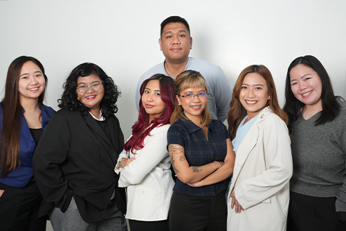 A diverse group of seven young professionals standing together against a light background, smiling confidently.