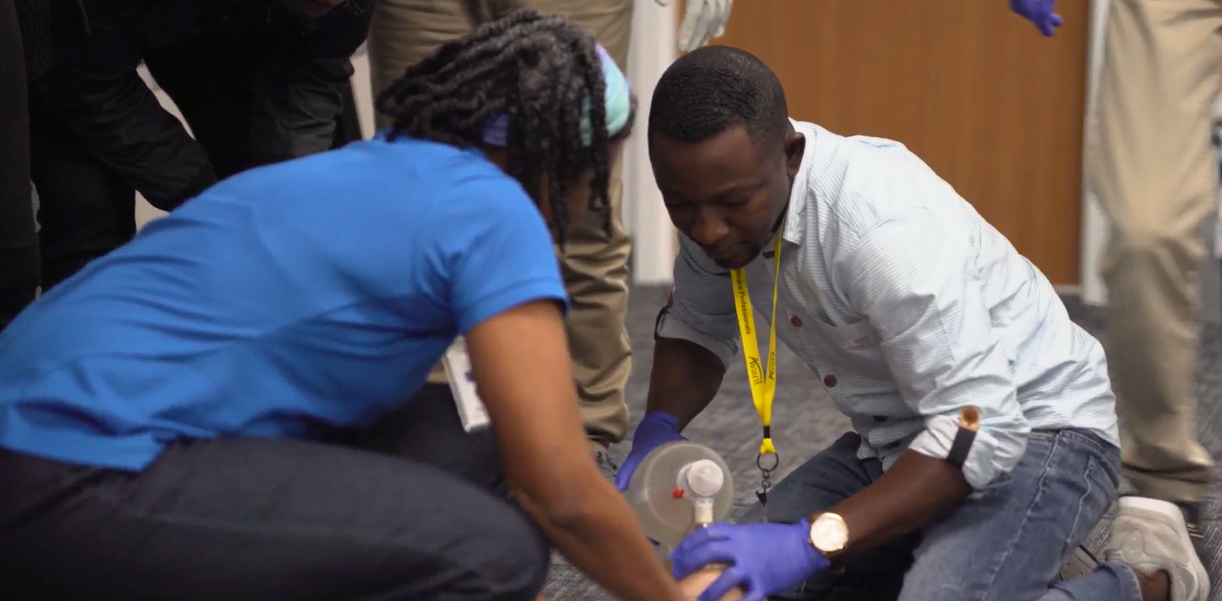 Two individuals in scrubs and casual clothes practice CPR techniques on a training mannequin.