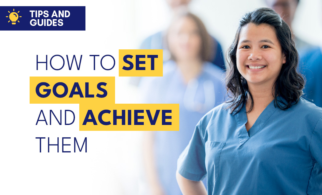 A smiling woman in medical scrubs stands confidently in a healthcare setting with blurred colleagues in the background.
